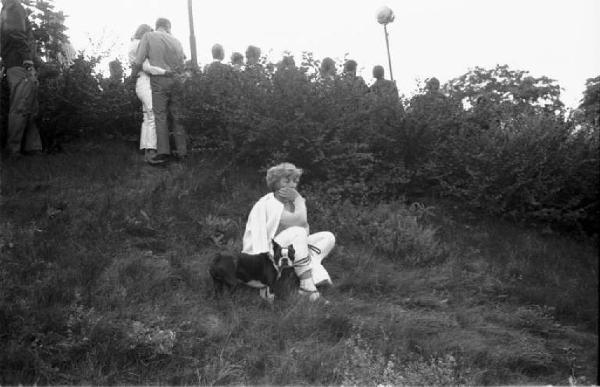 Svezia - Donna pensierosa seduta su di un pendio erboso - Sullo sfondo gruppo di gente voltato di spalle