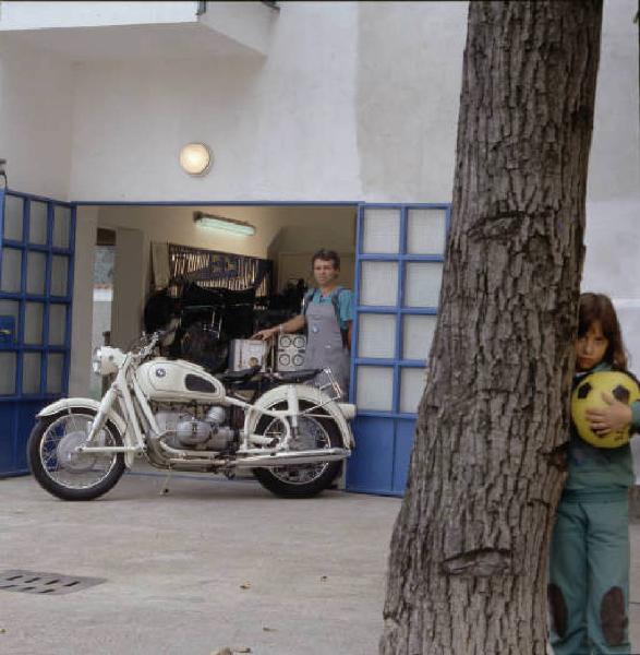 Officina di moto d'epoca - il titolare accanto a una motocicletta sull'ingresso dell'officina - sulla sinistra una bimba si nasconde dietro l'albero nel cortile