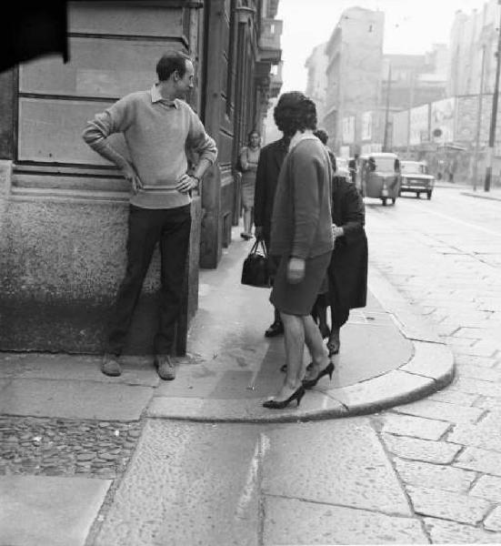 Milano - Giovane uomo con le mani sui fianchi in piedi sul marciapiede ad un angolo di strada guarda verso alcune donne che passeggiano
