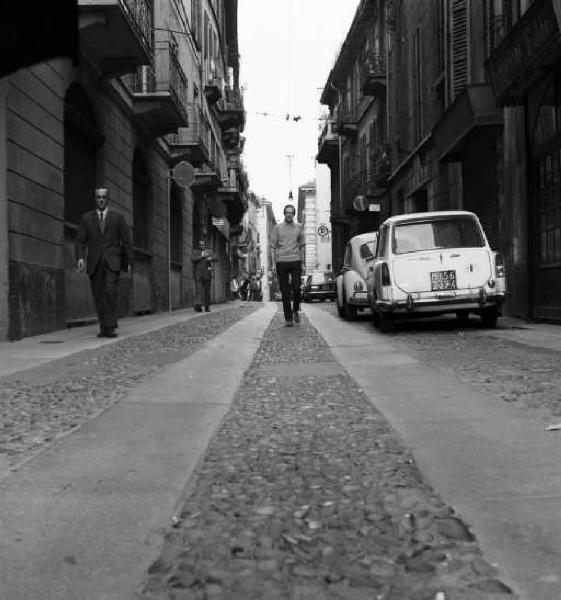 Milano - Giovane uomo passeggia al centro di una strada cittadina - auto parcheggiate e passanti