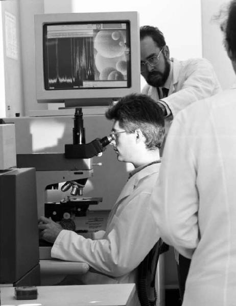 Glaxo - Ricercatore in camice bianco al microscopio - sullo sfondo uno schermo con encefalogramma - attorno altri medici