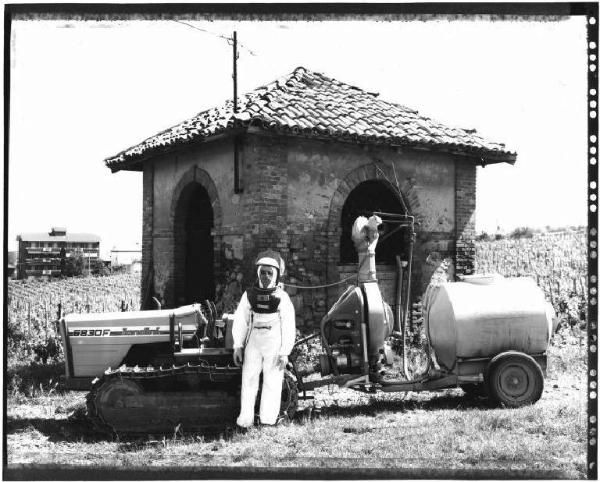 Uomo in tuta addetto ai diserbanti - trattore cingolato con traino-botte - casotto in mattoni - vigneto