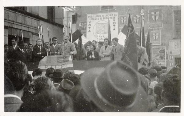 Casalpusterlengo - Elezioni politiche 1953 - Comizio del Partito Comunista Italiano - Francesco Scotti (a sinistra con le braccia incrociate) sul palco assiste al discorso di un oratore - Bandiere - Cartelli - Manifesti elettorali
