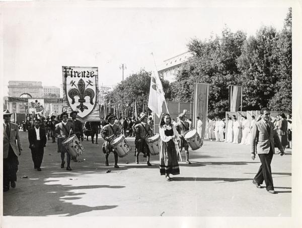 Genova - Festa nazionale de l'Unità 1950 - Sfilata per le vie della città - Spezzone di corteo con la rappresentanza della città di Firenze - Musicanti - Ragazza in costume - Bandiere - Cartelli - Striscione - Sullo sfondo l'Arco della Pace