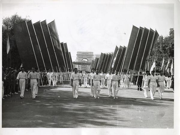 Genova - Festa nazionale de l'Unità 1950 - Sfilata per le vie della città - Spezzone di corteo con uomini vestiti di bianco che portano grandi bandiere montate su supporti di metallo - Striscione - Sullo sfondo l'Arco della Pace