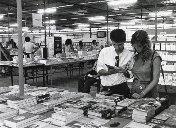 Reggio nell'Emilia - Festa nazionale de l'Unità 1983 - Lo stand della libreria Rinascita - Due giovani  osservano un libro - Illuminazione - Strutture metalliche - Cartelli - Ritratto di gruppo