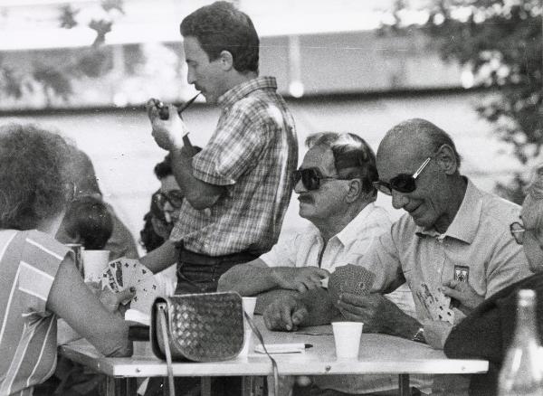 Milano - Festa provinciale de l'Unità 1984 - Monte Stella - Un gruppo di persone gioca a carte - Un uomo in piedi si accende la pipa - Ritratto di gruppo