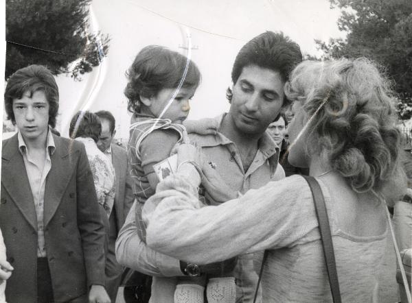 Roma(?) - Festa nazionale de l'Unità 1984(?) - Una "coccardista" mette l'adesivo sul vestito di un bimbo in braccio al papà - Ritratto di gruppo - Ritratto infantile