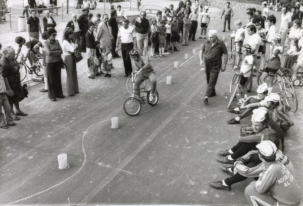 Bologna - Festa nazionale de l'Unità 1974 - Attività per i bambini - Gimkana in bicicletta - Giovane atleta in azione - Un uomo lo segue a piedi - Partecipanti seduti per terra - Spettatori - Ritratto