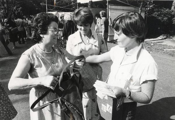 Milano - Festa nazionale de l'Unità 1979 - Parco Sempione - Una "coccardista" mette un adesivo sull'abito di una signora - Ritratto di gruppo - Stand