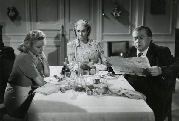 Scena del film "La famiglia Brambilla in vacanza" - Regia Carl Boese, 1941 - Attorno a una tavola imbandita, sulla destra Cesco Baseggio legge un giornale, al centro Amelia Chellini fissa Helen Lüber che ha lo sguardo rivolto verso il basso.

