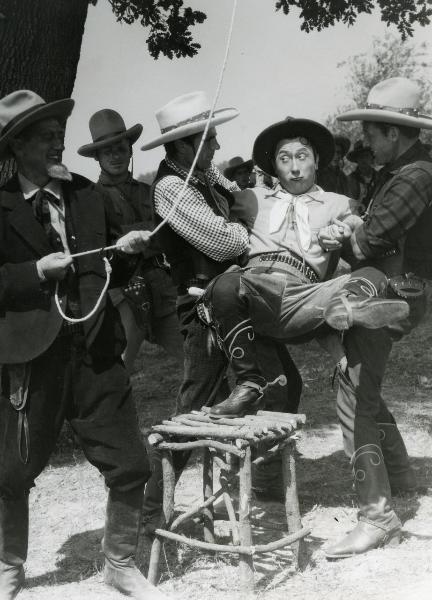 Scena del film "Il fanciullo del West" - Regia Ferroni, Giorgio, 1942 - Erminio Macario, in bilico su uno sgabello, si allontana da un attore non identificato a sinistra che tiene in mano un cappio. Intanto, è tenuto da due attori non identificati.