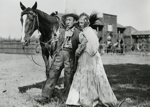 Scena del film "Il fanciullo del West" - Regia Ferroni, Giorgio, 1942 - Erminio Macario, al centro con in mano una pistola che guarda verso sinistra, e un'attrice non identificata che si nasconde dietro di lui e rivolge lo sguardo verso sinistra.