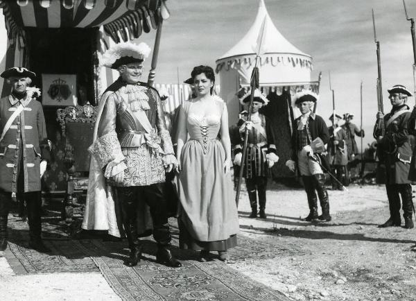 Scena del film "Fanfan la tulipe" - Regia Jacques, Christian, 1952 - Al centro, Marcel Herrand, in abiti eleganti, e Gina Lollobrigida, che rivolge lo sguardo verso sinistra, sono circondati da attori non identificati che indossano abiti militari.