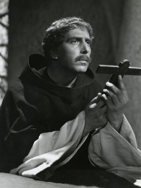 Scena del film "Fanfulla da Lodi" - Regia Duse, Carlo, Antamoro, Giulio, 1940 - Mezza figura di Ennio Cerlesi che, in abiti religiosi, regge tra le mani un crocifisso ligneo e rivolge lo sguardo davanti a sé.
