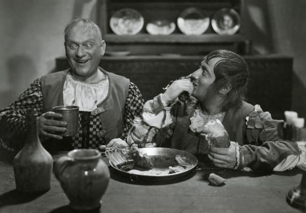 Scena del film "Fanfulla da Lodi" - Regia Duse, Carlo, Antamoro, Giulio, 1940 - Riccardo Billi, a destra, portandosi un pezzo di carne alla bocca, osserva Luigi Cantoni che guarda il bicchiere che regge nella mano destra.