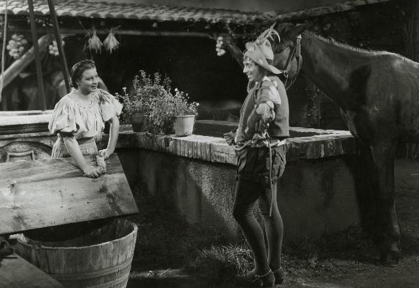 Scena del film "Fanfulla da Lodi" - Regia Duse, Carlo, Antamoro, Giulio, 1940 - Un attore non identificato, in piedi a destra, tiene le redini di un cavallo e guarda Marina Berti a sinistra che, lavando un panno, ricambia sorridendo.