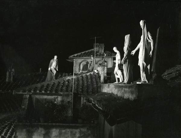 Scena del film "Fantasmi a Roma" - Regia Pietrangeli, Antonio, 1961 - Su un tetto, Marcello Mastroianni, a sinistra, osserva dritto davanti a sé, mentre un attore non identificato a destra legge. In primo piano, alcune statue classiche.