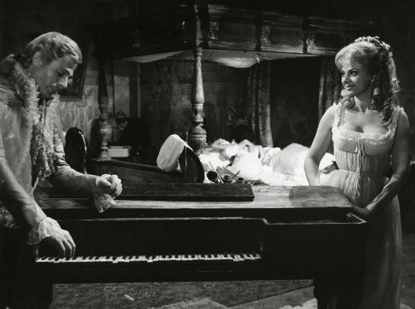 Scena del film "Fantasmi a Roma" - Regia Pietrangeli, Antonio, 1961 - In primo piano, Mastroianni appoggia la mano destra su un pianoforte e guarda in basso. Sandra Milo, a destra lo osserva. Dietro, distesi a letto, due attori non identificati.