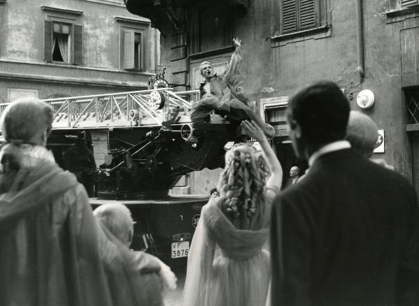 Scena del film "Fantasmi a Roma" - Regia Pietrangeli, Antonio, 1961 - Vittorio Gassman, seduto su un furgone dei pompieri, fa un cenno di saluto a degli attori non identificati in primo piano.
