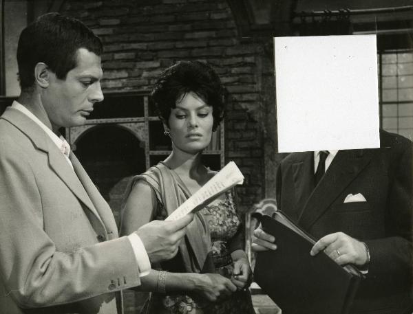 Scena del film "Fantasmi a Roma" - Regia Pietrangeli, Antonio, 1961 - Mastroianni, a sinistra, legge un documento. Intanto, Belinda Lee, al centro guarda in basso e un attore non identificato a destra tiene in mano un porta documenti.