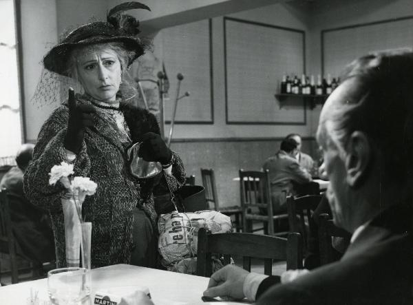 Scena del film "Fantasmi a Roma" - Regia Pietrangeli, Antonio, 1961 - Eduardo De Filippo seduto al tavolo di un ristorante, a destra, guarda un'attrice non identificata a sinistra che gli parla puntando l'indice della mano destra verso l'alto.
