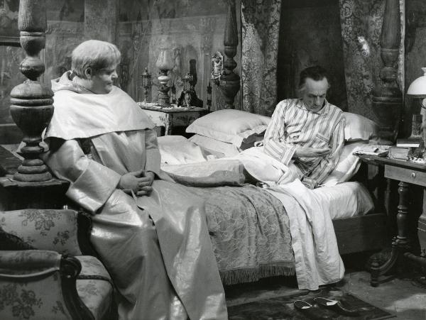 Scena del film "Fantasmi a Roma" - Regia Pietrangeli, Antonio, 1961 - Eduardo De Filippo, steso a letto, a destra, è intento a controllare l'orologio a cipolla che tiene in mano. Intanto, Tino Buazzelli, seduto ai piedi del letto, lo osserva.
