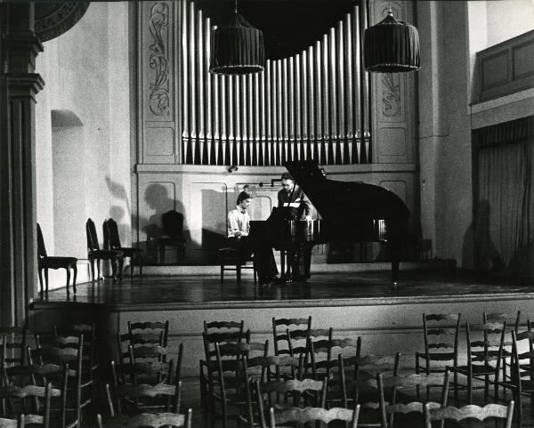 Scena del film "Una farfalla con le ali insanguinate" - Regia Tessari, Duccio, 1971 - Su un palcoscenico, Helmut Berger è intento a suonare un pianoforte a coda, mentre un attore non identificato, alla sua sinistra, lo osserva.