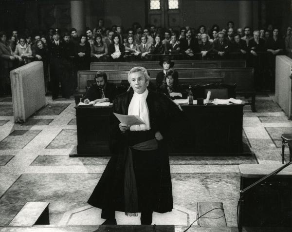 Scena del film "Una farfalla con le ali insanguinate" - Regia Tessari, Duccio, 1971 - In un'aula di tribunale: in primo piano, Gunther Stoll, in vesti di avvocato, mentre tiene in mano dei documenti e rivolge lo sguardo dritto davantio a sé.