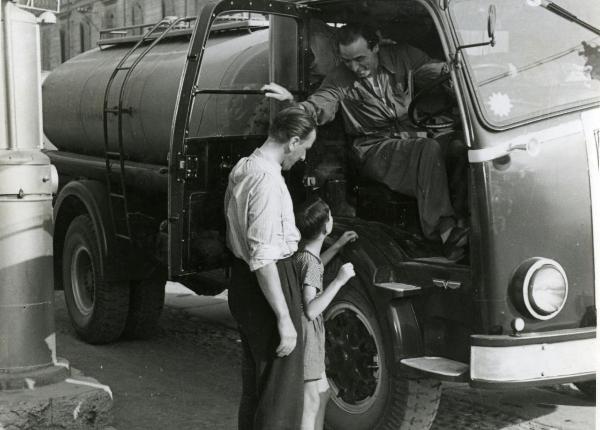 Scena del film "Fari nella nebbia" - Regia Franciolini, Gianni, 1942 - A destra, un attore non identificato, seduto sul sedile di un camion, si sporge verso l'esterno e conversa con un giovane attore non identificato appoggiato vicino alla ruota.