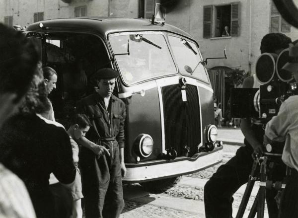 Sul set del film "Fari nella nebbia" - Regia Franciolini, Gianni, 1942 - A destra, un operatore non identificato rivolge la cinepresa verso un gruppo di attori non identificati a sinistra. Sullo sfondo è presente un camion.