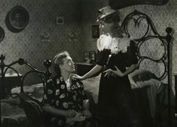 Scena del film "Fari nella nebbia" - Regia Franciolini, Gianni, 1942 - A sinistra, un'attrice non identificata seduta su un letto con i ferri da maglia, osserva un'attrice non identificata a destra che la guarda e le appoggia una mano sulla spalla.