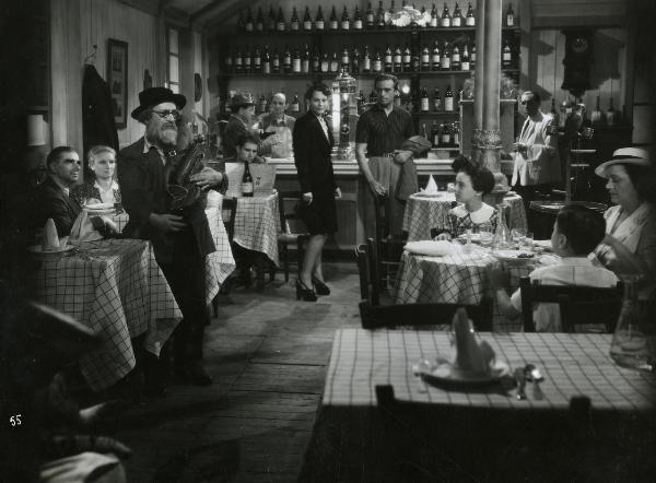 Scena del film "Fari nella nebbia" - Regia Franciolini, Gianni, 1942 - In un ristorante: Fosco Giachetti e Luisa Ferida, al centro, osservano un attore non identificato a sinistra, che tiene in mano un antico strumento musicale.