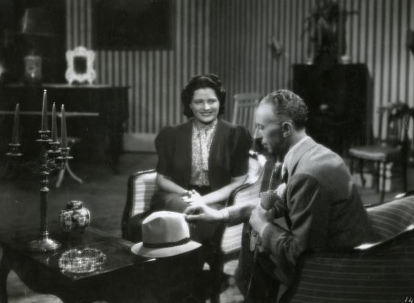 Scena del film "Fascino" - Regia Solito, Giacinto, 1939 - Seduti su due poltrone: Iva Pacetti, al centro, con le mani giunte, osserva sorridendo Giacomo Moschini, a destra, intento ad appoggiare un cappello su un tavolino antistante.