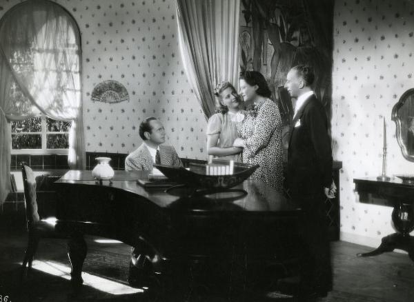 Scena del film "Fascino" - Regia Solito, Giacinto, 1939 - Cesare Bettarini, a sinistra seduto, osserva ricambiato Silvana Jachino, al centro che stringe la mano di Iva Pacetti. Questa la osserva come Giacomo Moschino, a destra.