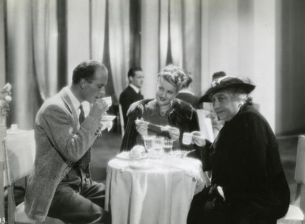 Scena del film "Fascino" - Regia Solito, Giacinto, 1939 - Iva Pacetti, al centro, con un coltello e una fetta di pane in mano, osserva Elio Stainer, a sinistra, mentre sorseggia da una tazza. A destra, Bella Starace Sainati, guarda alla sua destra.
