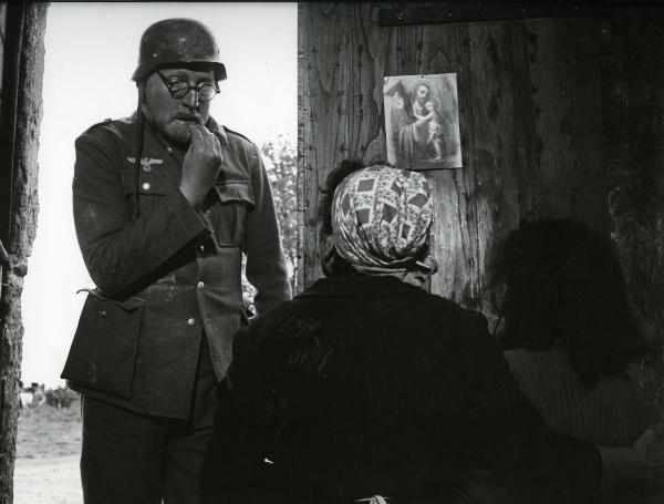 Scena del film "Il federale" - Regia Salce, Luciano, 1961 - Ugo Tognazzi, in primo piano con un elmo militare in capo, è seduto a braccia conserte e osserva in basso a sinistra. Alle sue spalle è incorniciato un ritratto fotografico del Duce.
