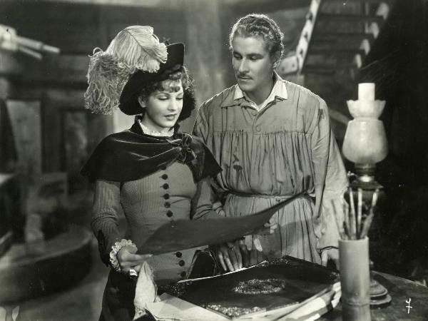 Scena del film "Fedora" - Regia Mastrocinque, Camillo, 1942 - Amedeo Nazzari, in veste di pittore, osserva Luisa Ferida, a sinistra, con un cappello decorato da una grande piuma che tiene in mano un grande foglio.