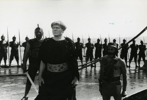 Scena del film "Fellini-Satyricon" - Fellini, Federico, 1969 - Su una spiaggia: al centro un attore non identificato, con l'occhio destro di vetro, guarda davanti a sé. In secondo piano, la schiera di attori non identificati, lo osservano.