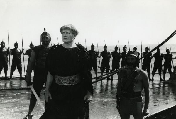 Scena del film "Fellini-Satyricon" - Fellini, Federico, 1969 - Su una spiaggia: al centro un attore non identificato, con l'occhio destro di vetro, guarda davanti a sé. In secondo piano, la schiera di attori non identificati, lo osservano.