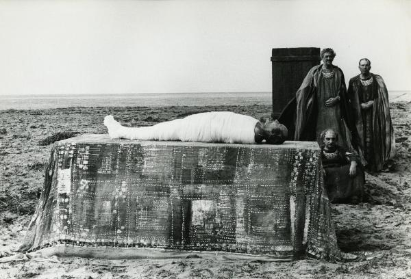 Scena del film "Fellini-Satyricon" - Fellini, Federico, 1969 - In secondo piano, due attori non identificati uno in piedi e uno seduto a terra osservano un attore non identificato in primo piano, steso supino su un altare, mummificato.
