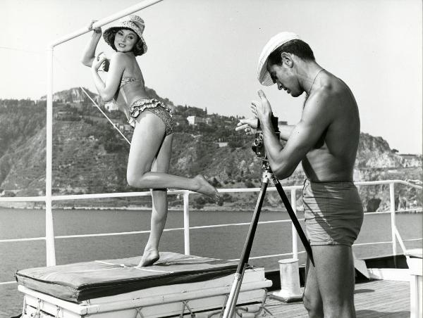 Scena del film "Femmine di lusso" - Bianchi, Giorgio, 1960 - Ugo Tognazzi, a sinistra, guarda in una fotocamera e dà indicazioni a Sylva Koschina che é in posa con un prodotto spray in mano.
