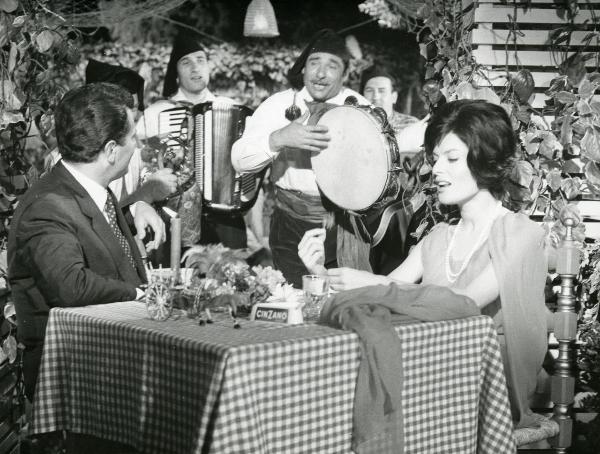 Scena del film "Femmine di lusso" - Bianchi, Giorgio, 1960 - Belinda Lee, a destra, guarda davanti a sé ed è seduta a un tavolo con Ugo Tognazzi che guarda in secondo piano, verso degli attori non identificati che suonano in abiti tradizionali.
