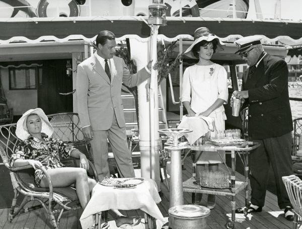 Scena del film "Femmine di lusso" - Bianchi, Giorgio, 1960 - A sinistra, un'attrice non identificata è seduta su una sedia di vimini e osserva davanti a sé. Al suo fianco Ivan Desny, in piedi, osserva Belinda Lee e un attore non identificato.