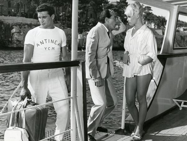 Scena del film "Femmine di lusso" - Bianchi, Giorgio, 1960 - Un attore non identificato tiene in mano delle valige e guarda verso sinistra. Intanto, a destra, un'attrice non identificata osserva Walter Chiari che si porta la mano alla fronte.