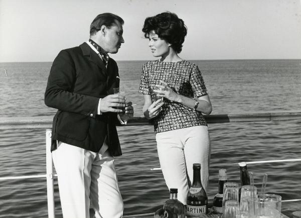 Scena del film "Femmine di lusso" - Bianchi, Giorgio, 1960 - Appoggiati alla balaustra di una nave, Ivan Desny, con una sigaretta in mano, e Belinda Lee conversano con due bicchieri in mano.
