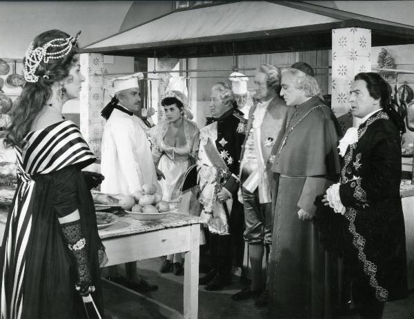 Scena del film "Ferdinando I, Re di Napoli" - Franciolini, Gianni, 1959 - Un'attrice non identificata, a sinistra, e un attore non identificato in vesti di cuoco, osservano alcuni attori tra cui è riconoscibile Vittorio De Sica, a destra.