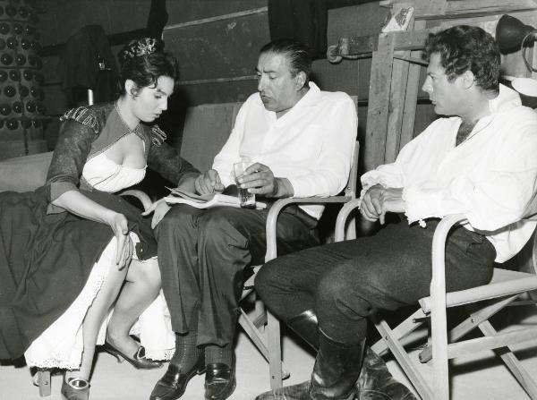 Sul set del film "Ferdinando I, Re di Napoli" - Franciolini, Gianni, 1959 - Marcello Mastroianni, seduto a destra, ascolta Jacqueline Sassard, a sinistra, e il regista Gianni Franciolini, al centro, che discutono osservando un copione.
