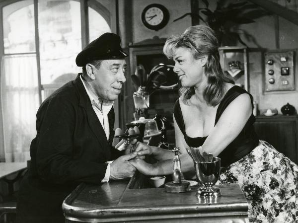 Scena del film "Fernandel, Scopa e Pennel" - Cloche, Maurice, 1961 - Fernandel osserva e stringe le mani di Dora Doll, dietro a un bancone, che ricambia sorridendo.
