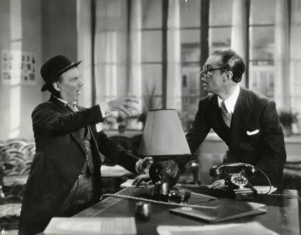 Scena del film "Il feroce Saladino" - Bonnard, Mario, 1937 - Intorno a una scrivania, un attore non identificato a destra, osserva un altro attore non identificato, a sinistra, che discute gesticolando.
